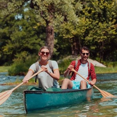 SUNGLASSES, BACKPACK, SHORTS, TREES, SMILING, COUPLE, CANOE, PADDLE, WATCH, RIVER, SHIRT